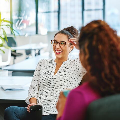 Zwei Frauen unterhalten sich lachend in einem modernen Büro mit grossen Fenstern und Pflanzen im Hintergrund.
