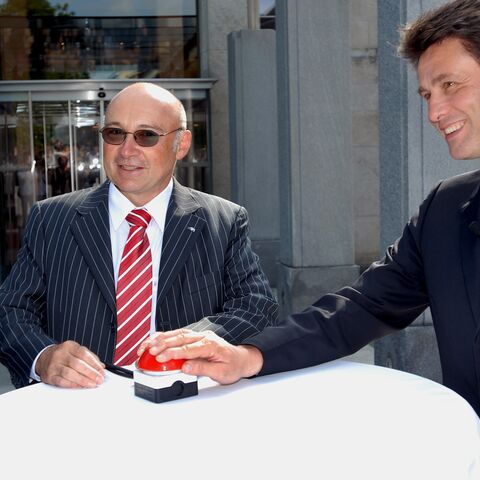 Two men in suits stand at a round table outside and press a big red button together.