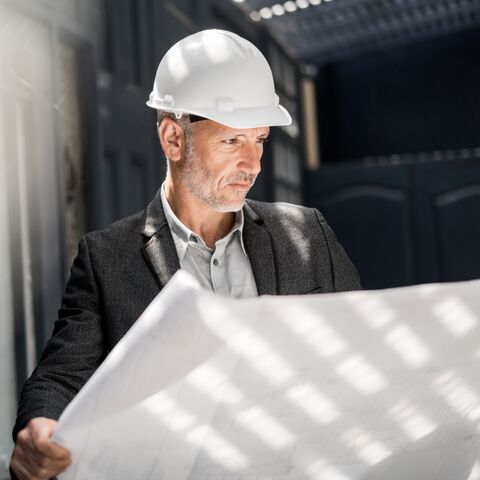 Un uomo di mezza età in giacca e cravatta e casco da cantiere osserva con attenzione un grande piano di costruzione in una stanza inondata di luce.