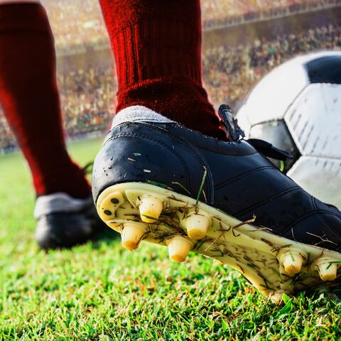 Un calciatore con il cappuccio rosso è in piedi su un campo verde e si prepara a tirare un pallone con la scarpa nera.