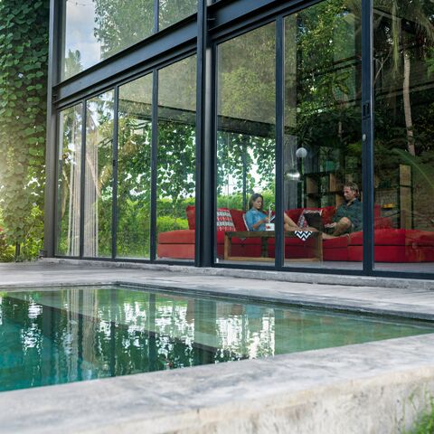 Ein modernes Haus mit grossen Glasfenstern, durch die ein Wohnzimmer mit rotem Sofa und zwei Personen sichtbar ist, davor ein Pool.