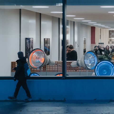 Im Vordergrund gehen in der Dämmerung zwei Personen auf einem Gehweg und schauen durch ein grosses Fenster, hinter dem man mehrere Personen bei der Vernissage einer Kunstausstellung sieht.