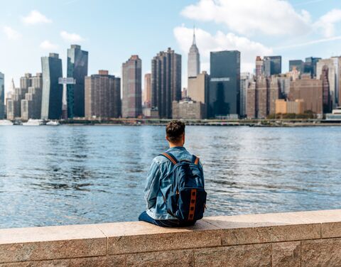 Ein Mann sitz in der USA, mit einem Rucksack auf dem Rücken vor einem Fluss und schaut auf Ihn und die Hochhäuser im Hintergrund. 