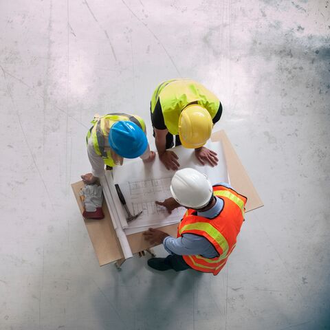 Trois ouvriers du bâtiment portant des équipements de sécurité et des casques discutent des plans de construction sur une table dans un chantier de construction en vue aérienne.