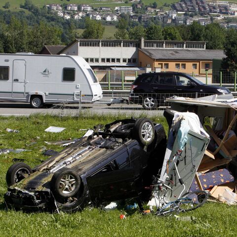 Ein umgestürztes Auto und ein zerstörter Wohnwagen auf einer Wiese neben einer Strasse mit vorbeifahrenden Fahrzeugen im Hintergrund.