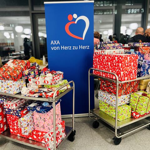 Two trolleys with colorfully wrapped gifts in front of an “AXA from heart to heart” banner in a busy room.