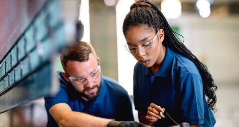 A young woman in blue work clothes and safety glasses holds a tablet while a man dressed the same way shows her something on the tablet.