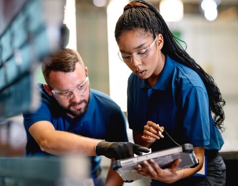 A young women in blue work clothes and protective glasses holds a tablet while a man dressed the same way shows her something on the tablet.