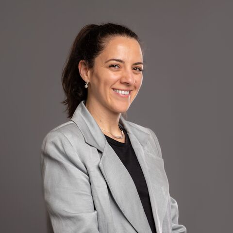 Eine junge Frau mit langen, zu einem Pferdeschwanz zusammengebundenen Haare lächelt freundlich in die Kamera. Sie trägt einen graublauen Blazer und ein schwarzes V-Neck darunter.