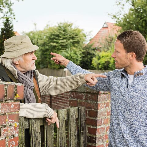 Un homme plus âgé et un homme plus jeune se tiennent devant un mur de briques et se montrent en gesticulant alors qu’ils se disputent apparemment.