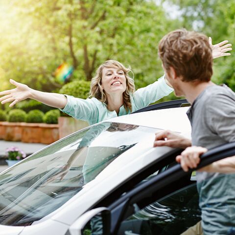 Eine Frau mit ausgebreiteten Armen begrüsst freudig einen Mann, der aus einem weissen Auto steigt, vor einem grünen, sonnigen Hintergrund.