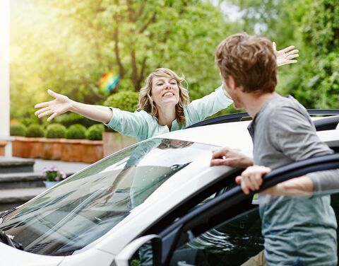 Una donna con le braccia tese saluta con gioia un uomo che scende da un'auto bianca su uno sfondo verde e soleggiato.