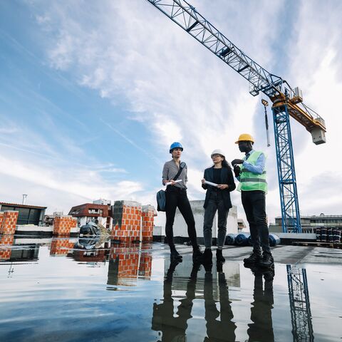Three construction workers in protective gear discuss on a building site with a large crane in the background and reflect in a puddle of water.