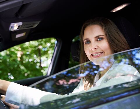 Eine Frau mit langen braunen Haaren lächelt, während sie in einem Auto sitzt und durch das offene Fenster auf der Fahrerseite schaut.