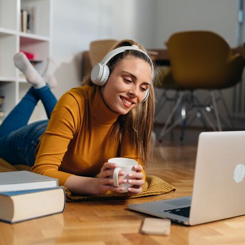 Eine Frau liegt auf dem Boden ihres Wohnzimmers, lächelt und schaut auf ihren Laptop.
