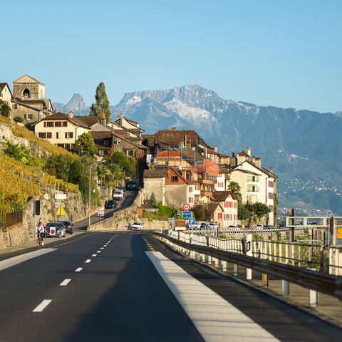 Una strada di campagna conduce attraverso un pittoresco villaggio con vigneti e case tradizionali, circondato da montagne sotto un cielo limpido.