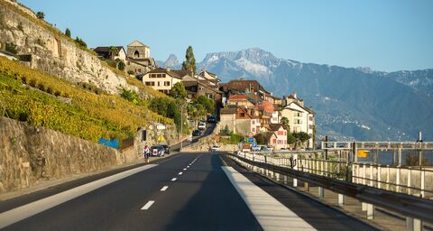 Una strada di campagna conduce attraverso un pittoresco villaggio con vigneti e case tradizionali, circondato da montagne sotto un cielo limpido.
