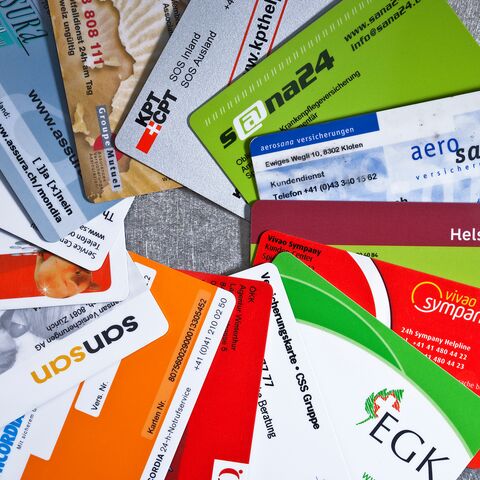 Verschiedene Krankenkassenkarten liegen verstreut auf einer grauen Oberfläche, darunter Karten von Sanitas, Helsana und anderen Anbietern. 