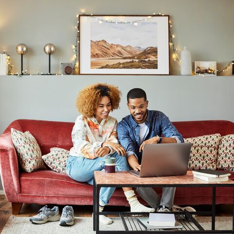 Un jeune couple est assis sur le canapé dans le salon de la maison et tous deux regardent joyeusement leur ordinateur portable.