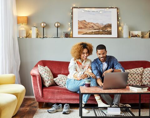 A young couple is sitting at home on the sofa in the living room and both are looking happily at the laptop.