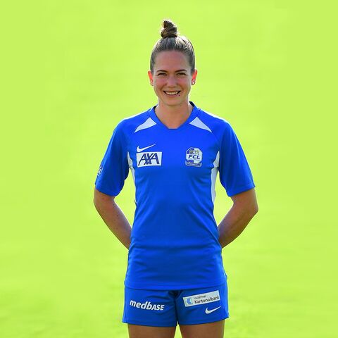 Eine lächelnde Frau in einem blauen Sporttrikot steht vor einem grünen Hintergrund mit den Händen hinter ihrem Rücken.