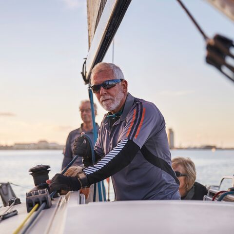 Ein älterer Mann trägt Sonnenbrille und steuert ein Segelboot bei Sonnenuntergang, begleitet von zwei weiteren Personen an Bord.