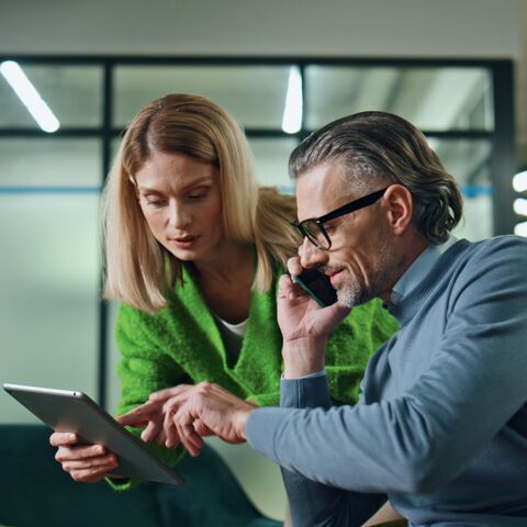 A women in a green sweater shows something on a tablet to a man in glasses on the phone.