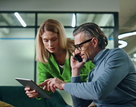 A women in a green sweater shows something on a tablet to a man in glasses on the phone.