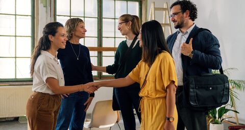 Five people are chatting amicably in a bright room; two women are shaking hands while the others smile and watch.