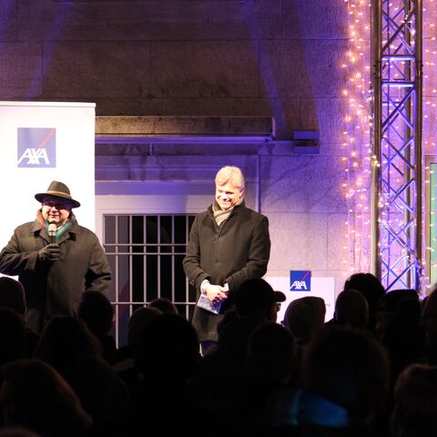 Two men stand on a stage in front of an audience; the stage is festively decorated with lights and an AXA logo can be seen.