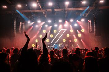 Scena di concerto con palco illuminato a colori e pubblico festante in primo piano.