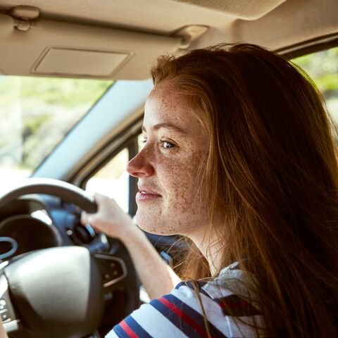 Une femme avec des taches de rousseur est au volant d’une voiture et roule sur une route ensoleillée avec un arrière-plan flou.