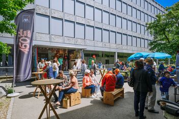 People sit and stand at outdoor tables and benches in front of the Flora Futura Box. A flag with the inscription “150 years of AXA” marks the venue.