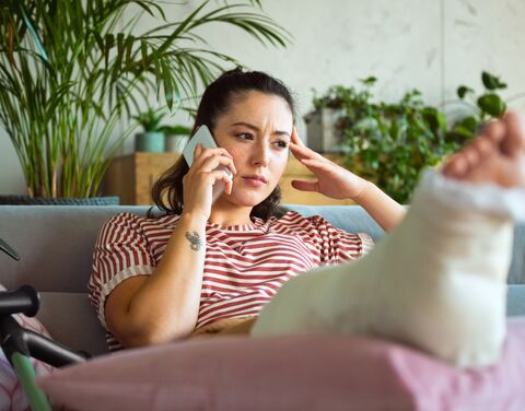 A woman with a plastered leg sits on a sofa talking on the phone, surrounded by plants in the background. She looks worried.