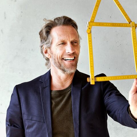 A smiling man in a dark jacket holds a yellow folding rule house model in front of a plain, light-colored background.