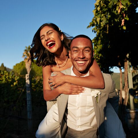 Un couple souriant, la femme étreint l’homme par derrière, tous deux élégamment habillés et debout sur un fond vert et ensoleillé.
