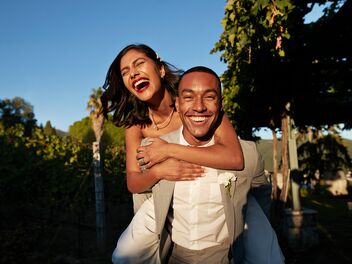 Un couple souriant, la femme étreint l’homme par derrière, tous deux élégamment habillés et debout sur un fond vert et ensoleillé.