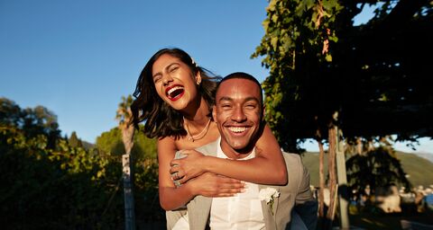 Una coppia sorridente, la donna abbraccia l’uomo da dietro, entrambi elegantemente vestiti e in piedi su uno sfondo verde e soleggiato.