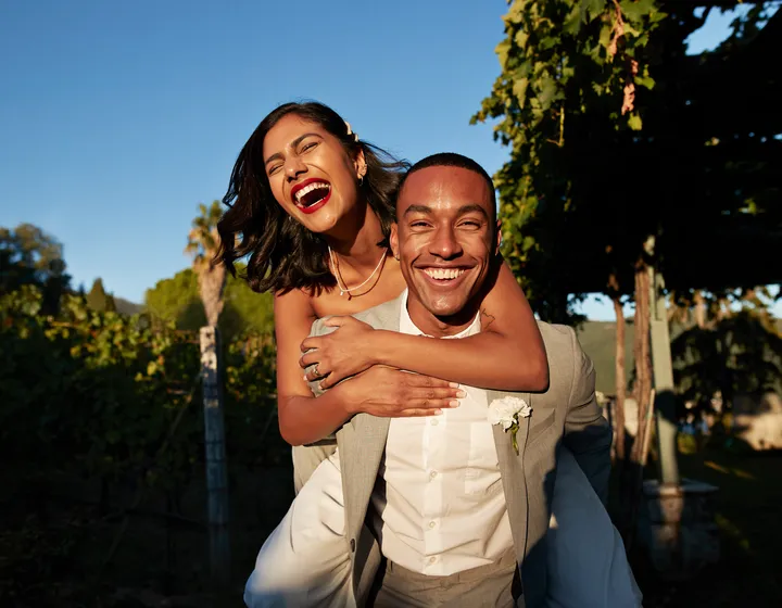 Una coppia sorridente, la donna abbraccia l’uomo da dietro, entrambi elegantemente vestiti e in piedi su uno sfondo verde e soleggiato.