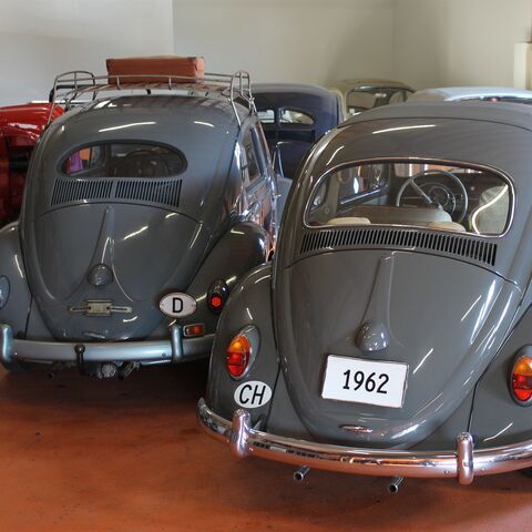 Due maggiolini VW grigi, uno dei quali con la targa “1962”, sono parcheggiati in una sala con altre auto d'epoca su un pavimento rosso.