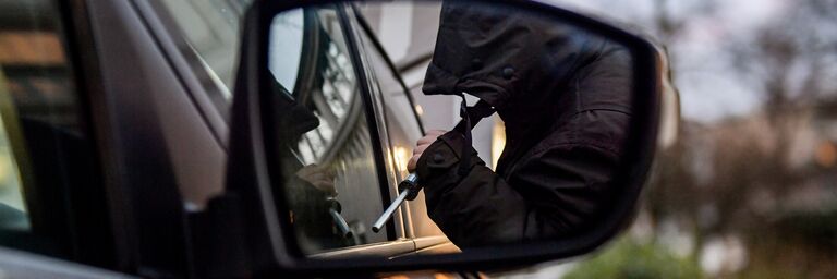 Dans le rétroviseur latéral d'une voiture, on aperçoit un homme cagoulé qui tente de forcer la voiture