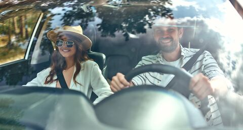Ein glückliches Paar geniesst eine Autofahrt durch einen Wald, gesehen durch die spiegelnde Windschutzscheibe.