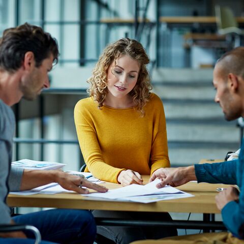 Trois personnes sont assises à une table dans un bureau moderne, discutent de documents et travaillent ensemble sur un projet.