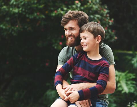 Ein Vater sitzt im Grünen mit seinem Sohn auf dem Schoss.