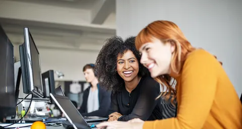Zwei Frauen arbeiten am Computer im Büro und lachen dabei.