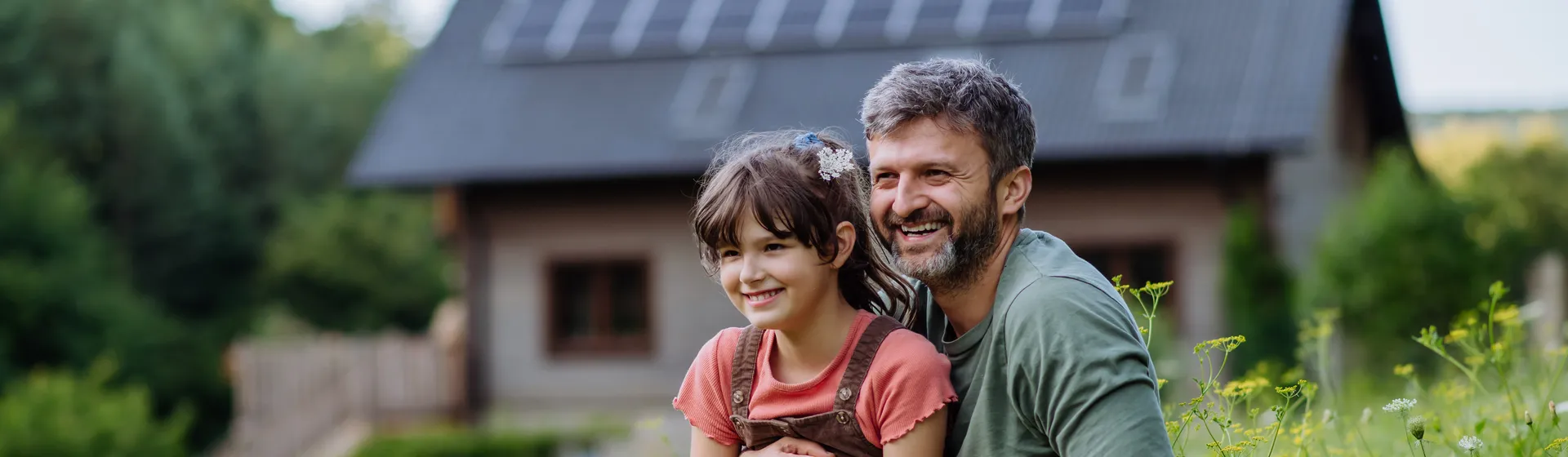 Un padre è seduto con la figlia su un prato fiorito ed entrambi guardano in lontananza con un sorriso. Sullo sfondo si trova la loro casa.