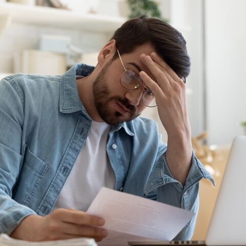 Ein Mann sitzt vor seinem Laptop, fasst sich an die Stirn und hält ein Blatt in der Hand.
