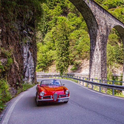 Un cabriolet rouge roule sous un vieux pont de pierre sur une route sinueuse qui traverse un paysage verdoyant et boisé.