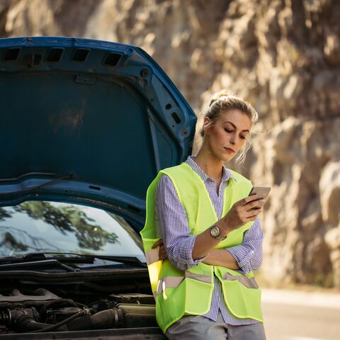 Una donna con un giubbino catarifrangente guarda nel suo cellulare e aspetta il soccorso stradale. Si appoggia alla sua auto nera.