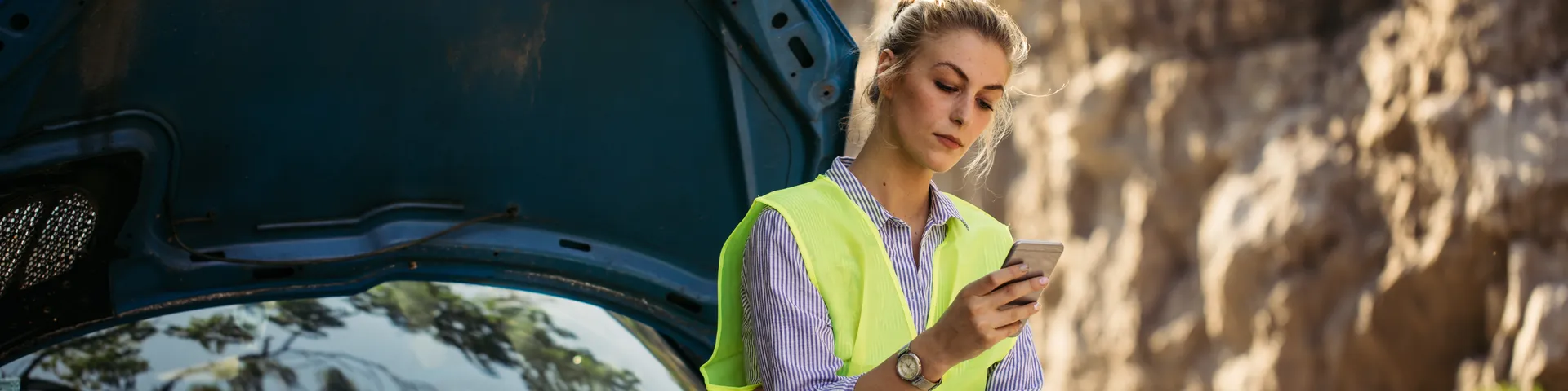 Eine Frau mit Leuchtweste schaut in ihr Handy und wartet auf die Pannenhilfe. Dabei lehnt sie sich an ihrem schwarzen Auto an.